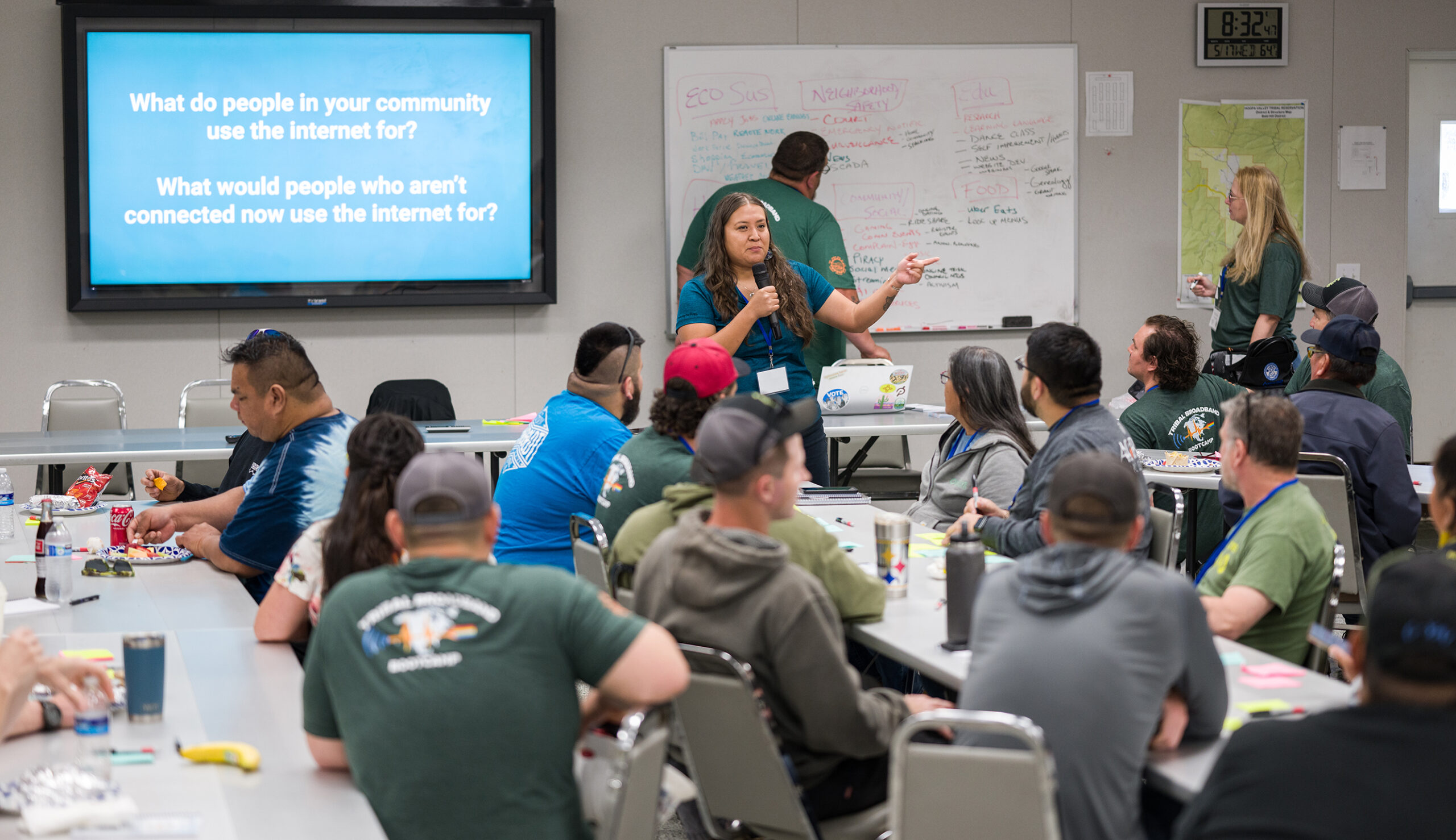 A conference room with a presentation showing a blue slide with white text in the background, and a group of people in casual clothing sitting at tables in the foreground. Davida Delmar stands in front of the presentation and speaks into a microphone.