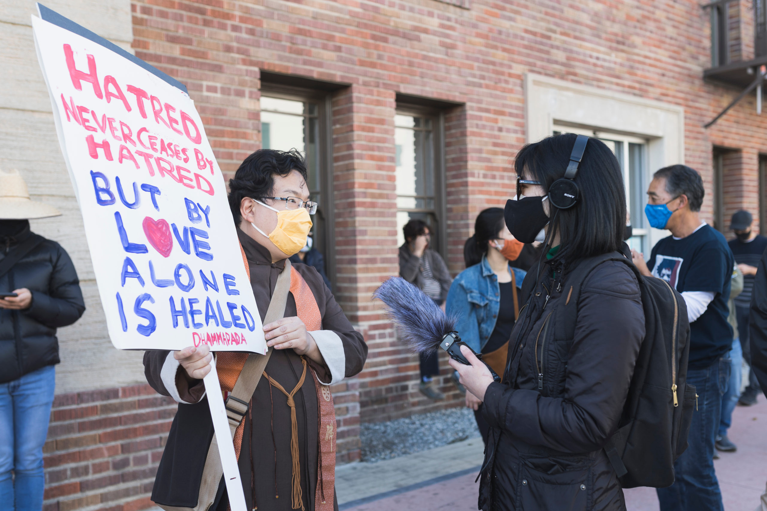 Reporter interviews a protester with a sign saying "Hatred never ceased by hatred but by love alone is healed -Dhammapada"